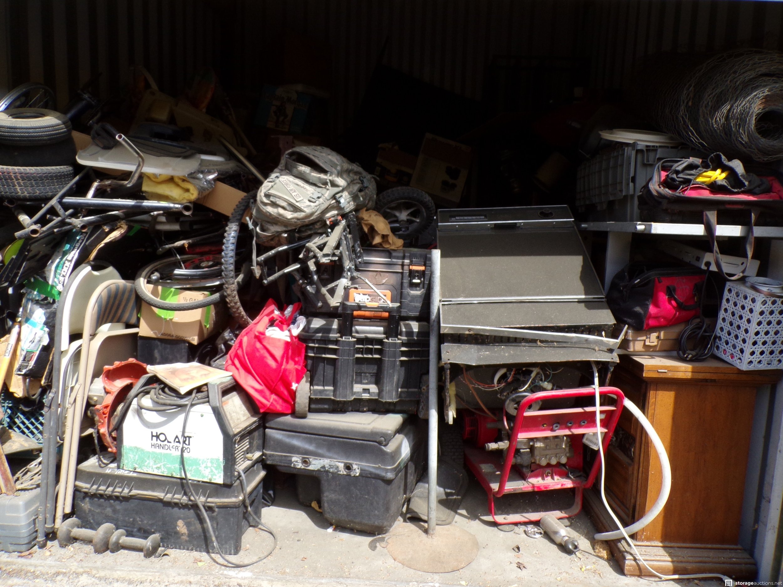 Cluttered garage or storage space with various tools, equipment, and household items, including a red hand truck, black tool boxes, cardboard boxes, and a wooden cabinet.