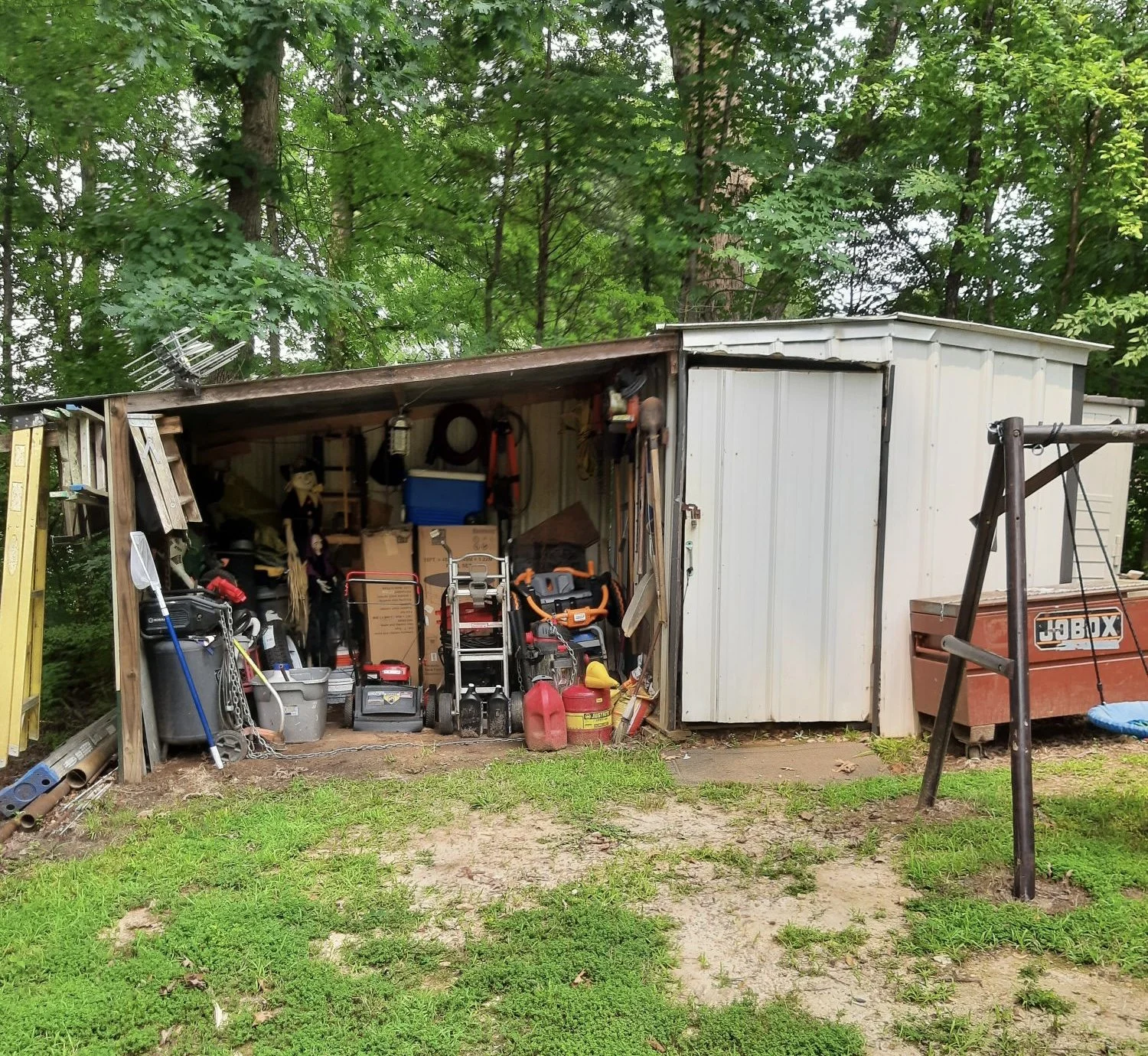 Backyard shed filled with various gardening and yard tools, lawnmower, chainsaw, and storage boxes, with a swing set frame nearby, surrounded by green trees.