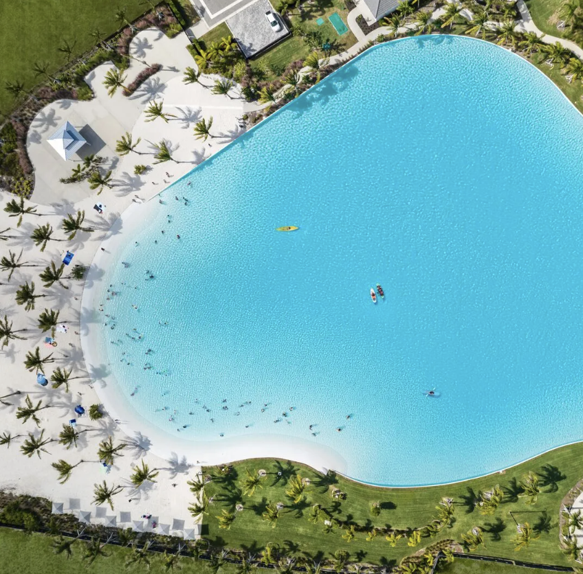 Aerial view of a large, curved swimming pool surrounded by palm trees, sandy area with umbrellas, and green grass.