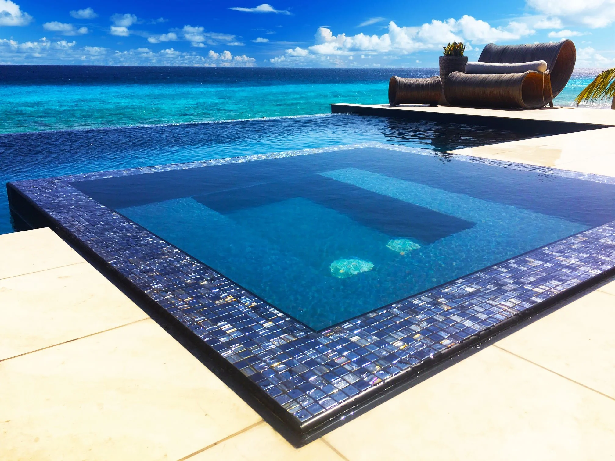 An infinity pool overlooking the ocean with a lounge chair and a small side table on a deck, under a blue sky with clouds.