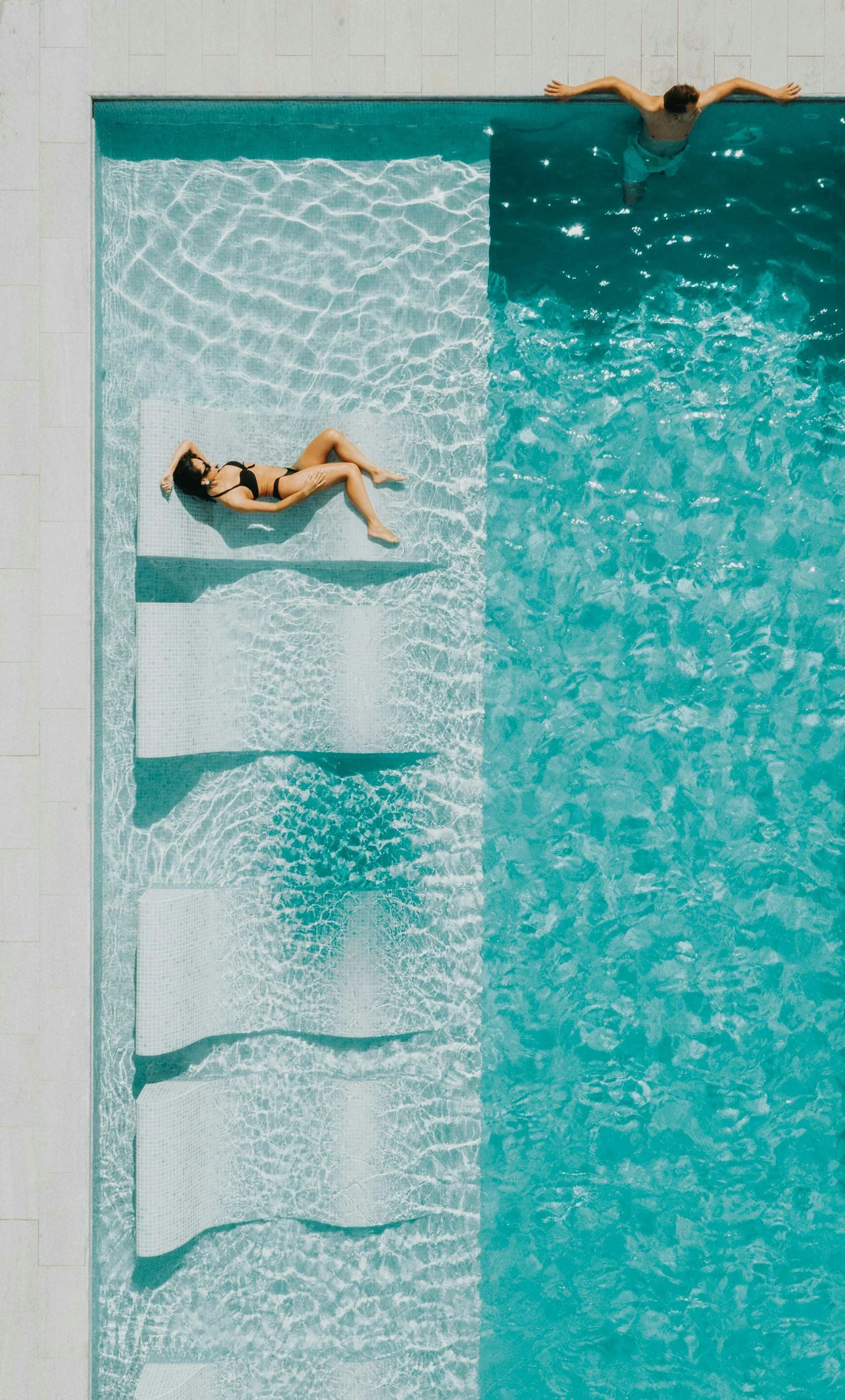 Overhead view of a swimming pool with a woman in a black bikini laying on a pool float on the left and a man in swim trunks swimming on the right.