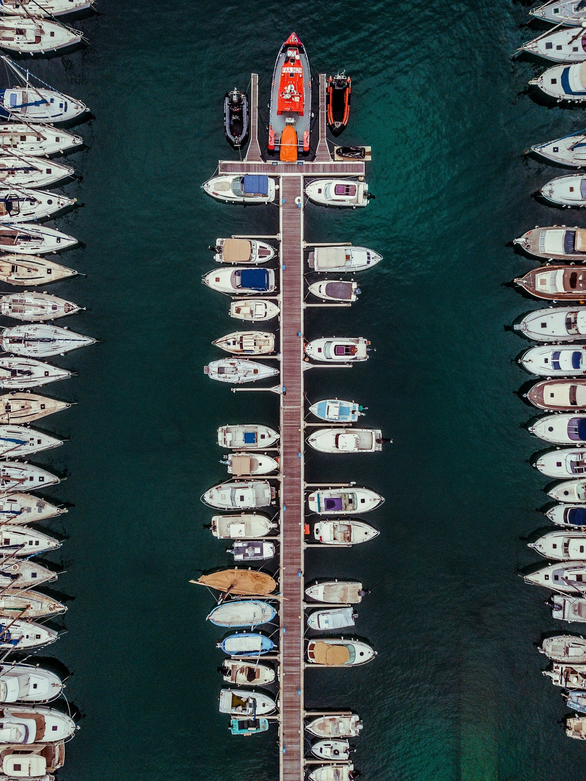 An aerial view of a marina with a wooden dock extending into the water, and numerous boats and yachts moored on either side.
