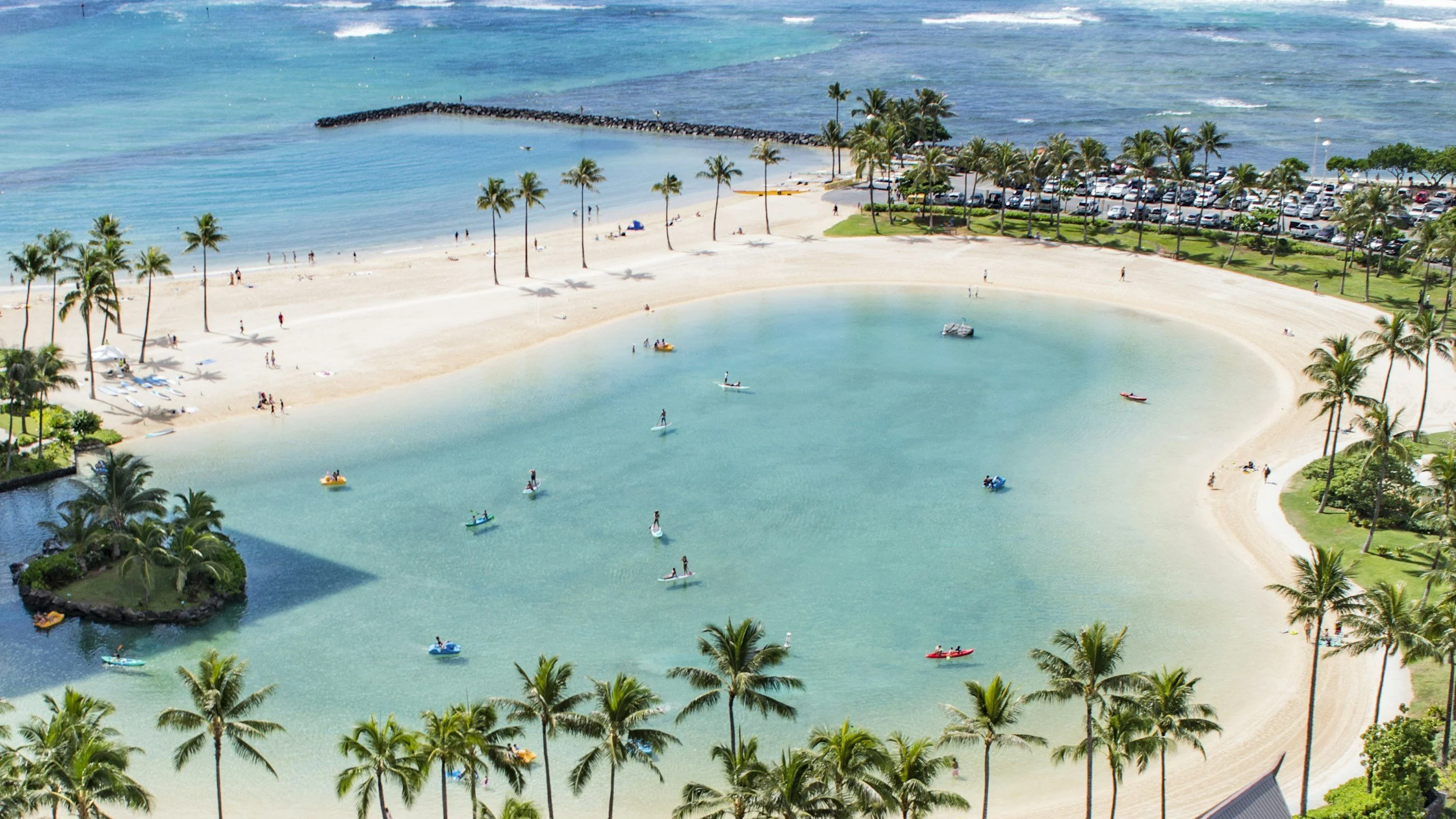 A scenic view of a tropical beach with calm, turquoise water, surrounded by palm trees and a sandy shoreline. People are engaging in paddleboarding and relaxing by the water, with a parking lot and ocean visible in the background.