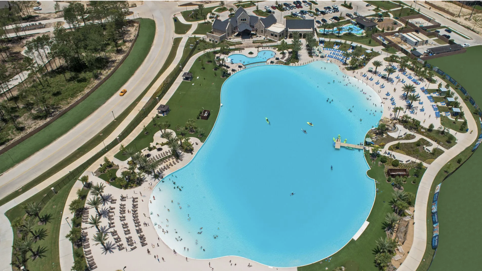 Aerial view of a large community swimming pool surrounded by lounge chairs, palm trees, and landscaped areas. There are several people swimming and floating in the pool, with a smaller kiddie pool nearby. The area has pathways, shaded seating spots, and nearby buildings, with a parking lot and wooded area in the background.
