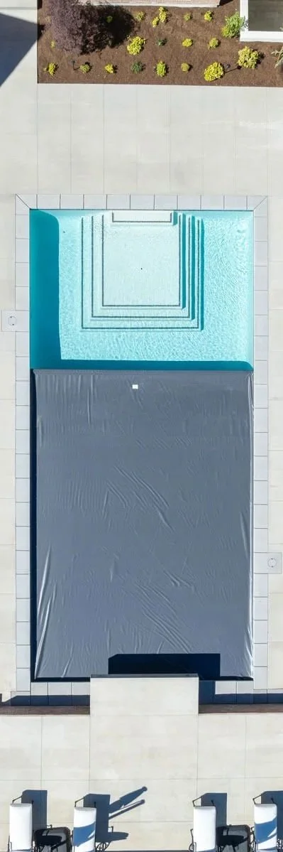 A backyard with a swimming pool that has steps, a cover on part of the pool, and a tiled deck area with lounge chairs. A garden bed with plants and shrubs is visible at the top.