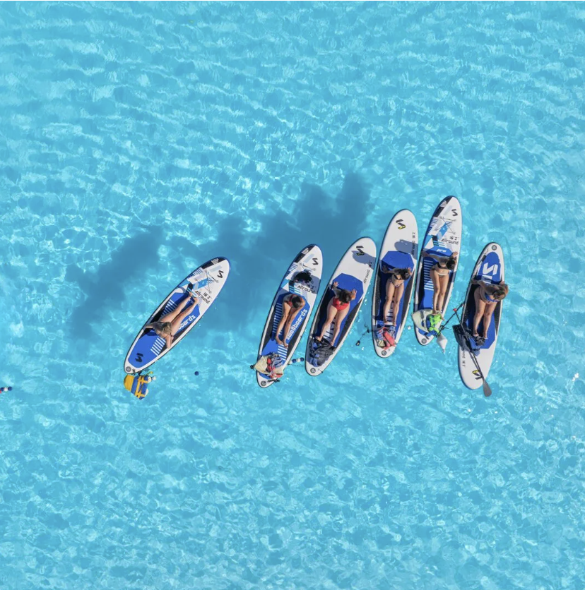 An aerial view of seven people on stand-up paddleboards in bright blue water, some sitting and some standing, with a few carrying paddles and belongings.