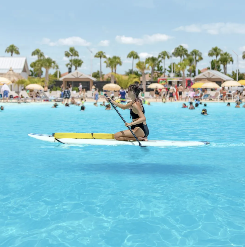 A woman sitting on a paddleboard in a pool, holding a paddle, with a busy poolside area and palm trees in the background.