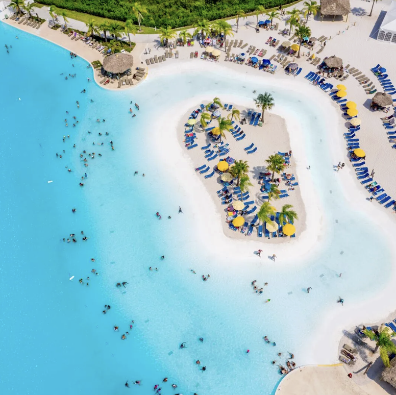 Aerial view of a large resort swimming pool filled with people, surrounded by lounge chairs, umbrellas, and palm trees, with a sandy area and greenery nearby.