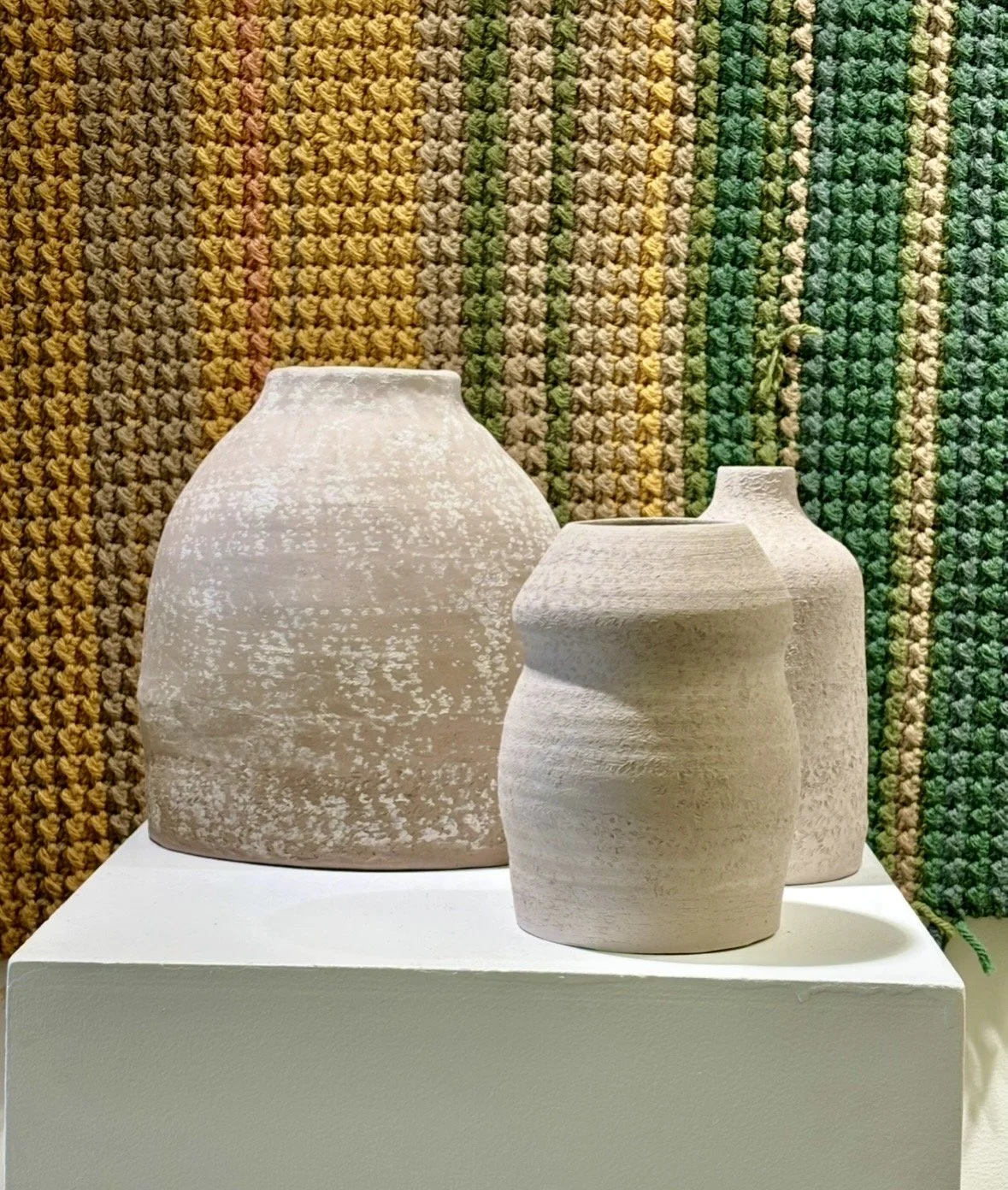 Three textured, off-white ceramic vases placed on a white platform against a multicolored, woven, striped background.