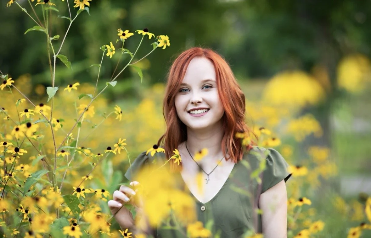 A young woman with red hair smiling in a field of yellow flowers.