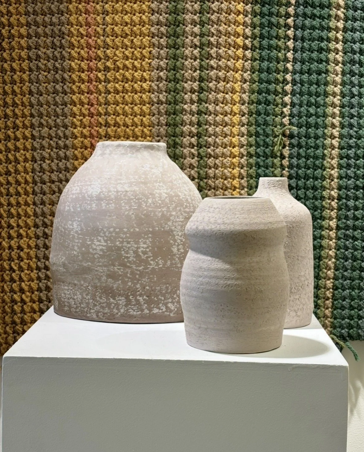 Three white ceramic vases with textured surfaces on a white pedestal, with a multicolored knit wall in the background.