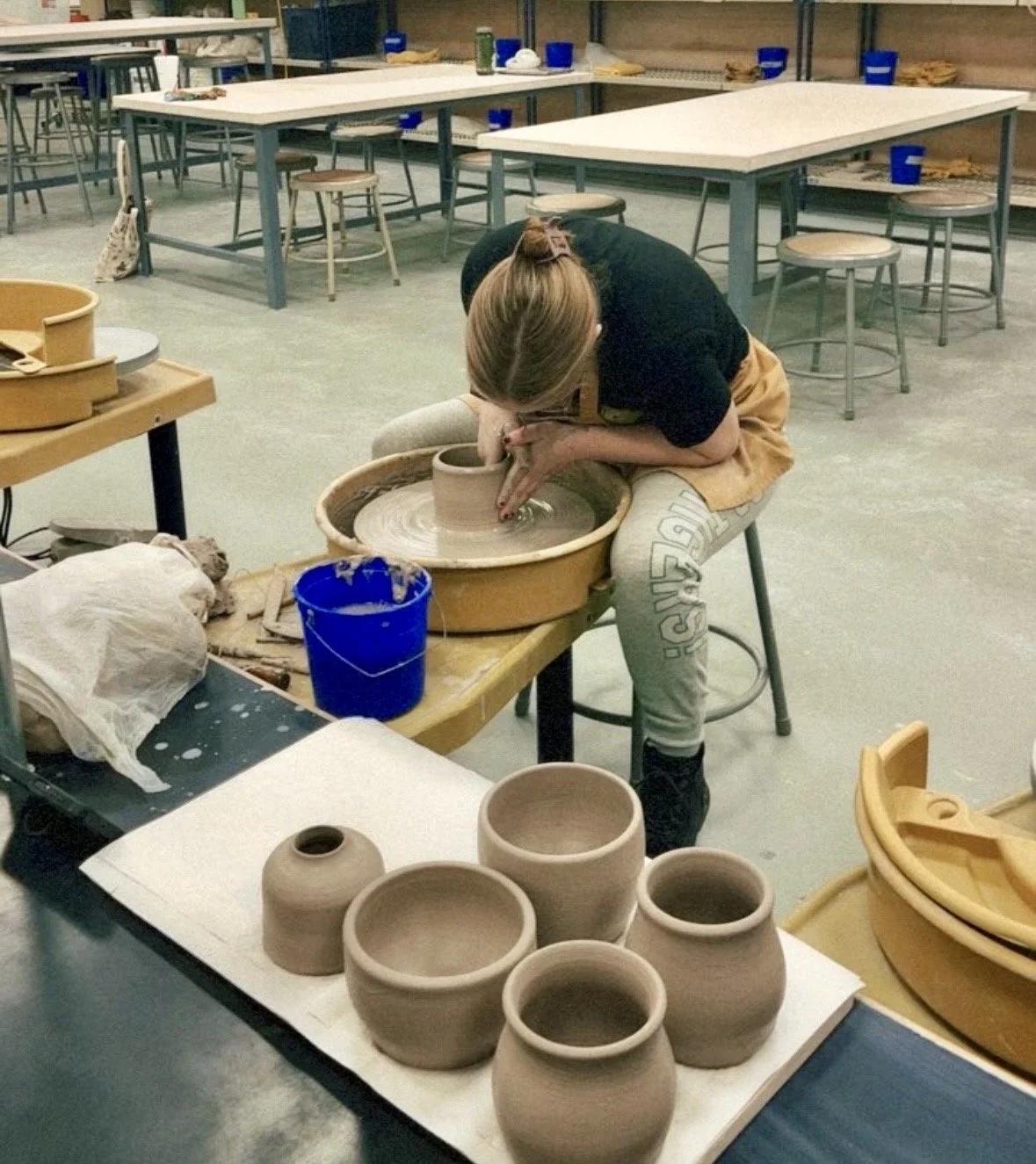 A person working on pottery at a pottery wheel in a workshop, surrounded by several finished clay pots, with tables and chairs in the background.