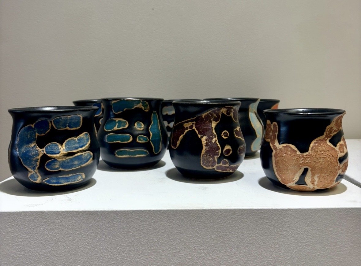 Six black ceramic pots with multicolored gold and turquoise abstract patterns displayed on a white pedestal against a plain wall.