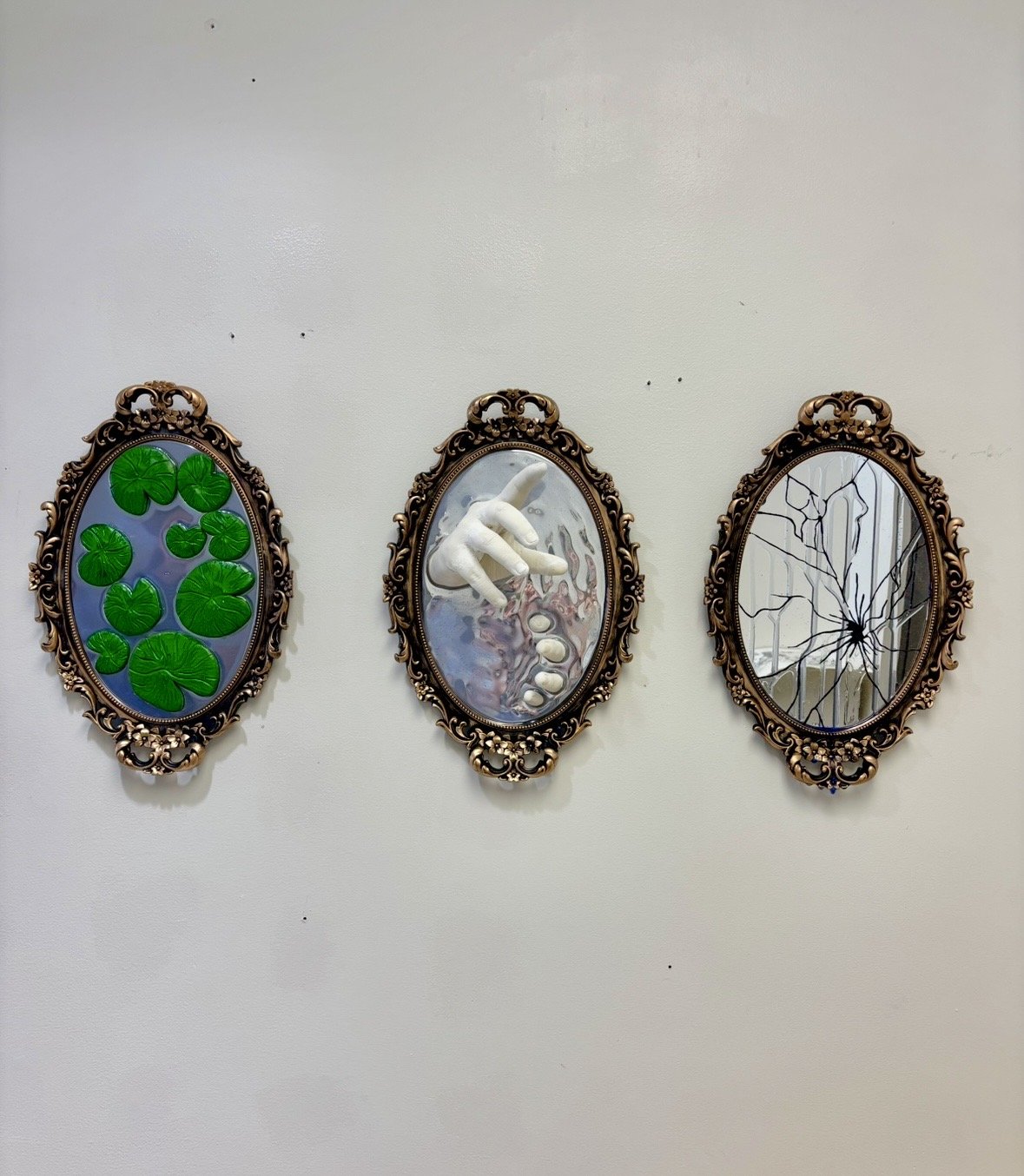 Three ornate oval mirrors on a wall, each with different artwork reflection: the left mirror shows green leaves, the middle shows a white hand with rings, and the right shows a broken glass pattern.