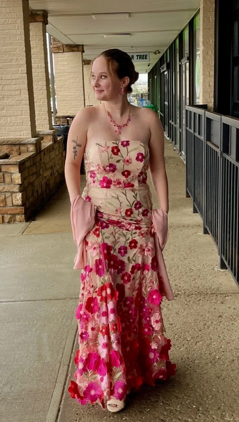 A young woman in a strapless floral dress with pink, red, and purple flowers, standing on a sidewalk outside a building with brick and glass walls, smiling and looking to her side.