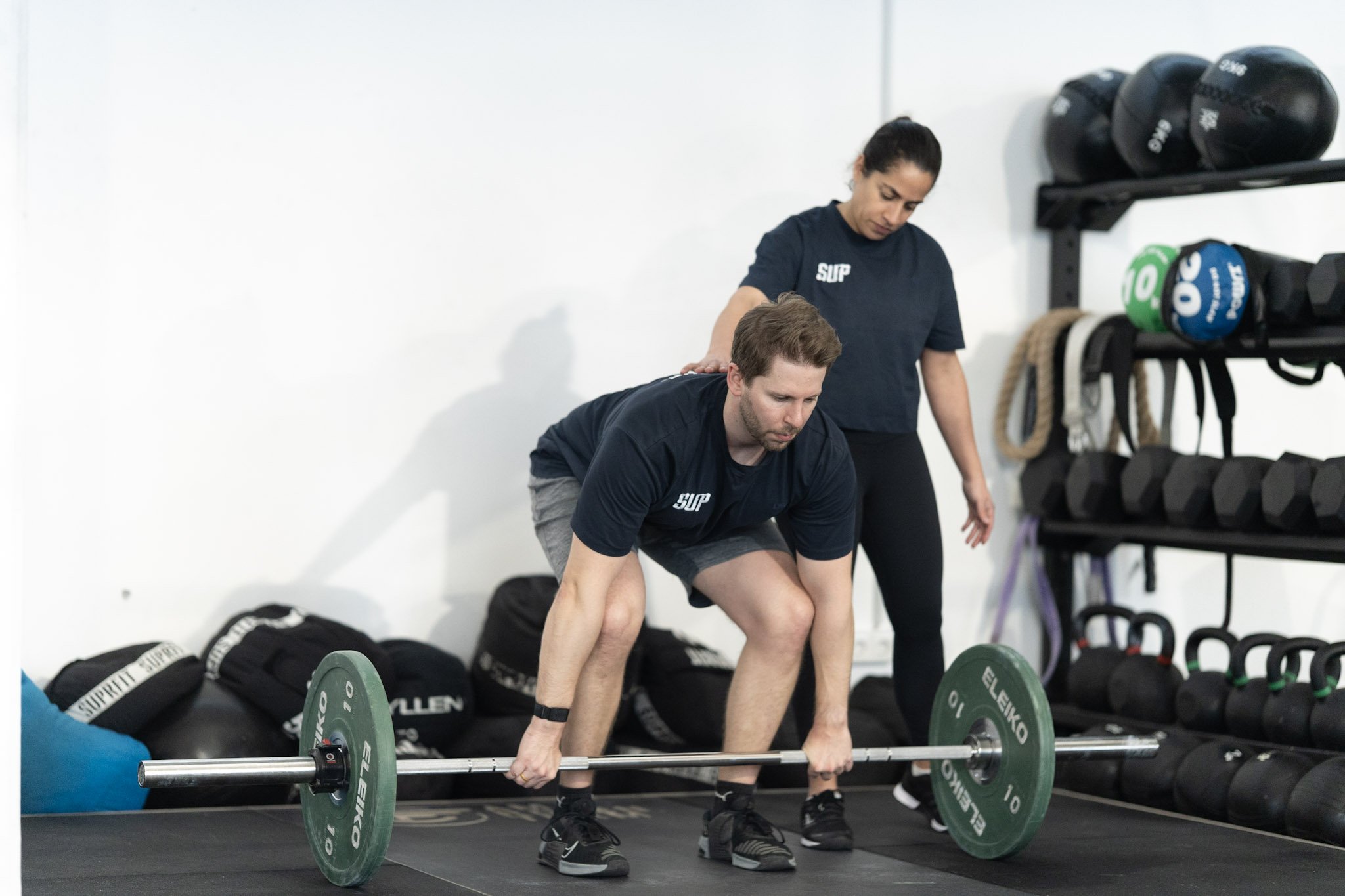 Eine Frau bei einer Krafttrainingseinheit im Fitnessstudio hebt eine Langhantel, während eine Trainerin neben ihr sie korrigiert. Im Hintergrund sind Hanteln und Fitnessgeräte sichtbar.