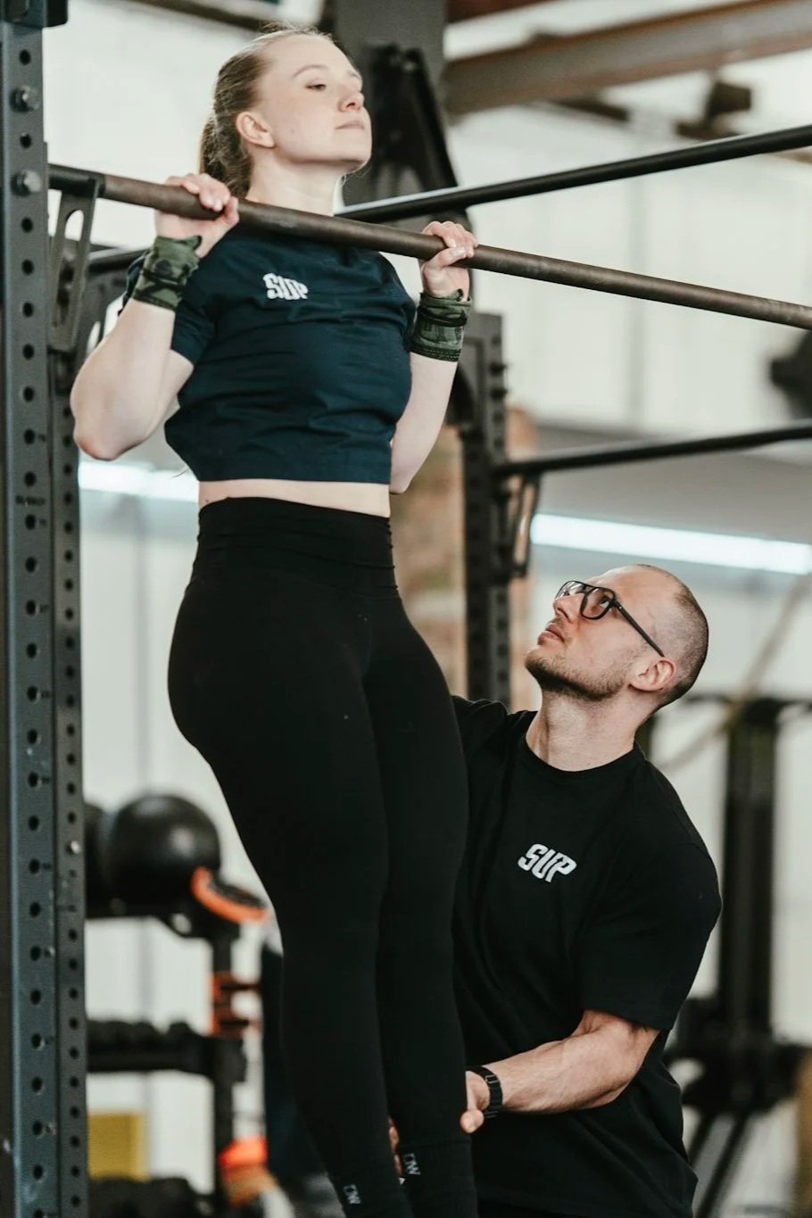Frau macht Kniebeugen mit einem Langhantel, während ein Trainer im Fitnessstudio neben ihr steht und sie unterstützt.