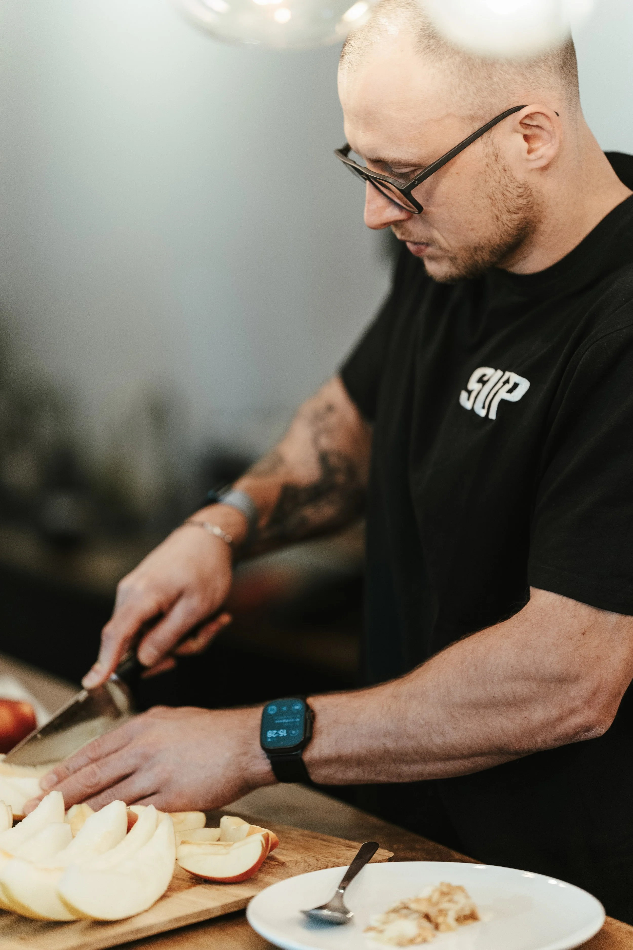 Ein Mann schneidet mit einem Messer auf einem Schneidebrett mit geschnittenen Apfelscheiben. Er trägt eine schwarze Brille, ein schwarzes T-Shirt mit weißen Buchstaben und eine Smartwatch am Handgelenk. Im Vordergrund befindet sich ein Teller mit einem Löffel und einem Stück Kuchen.