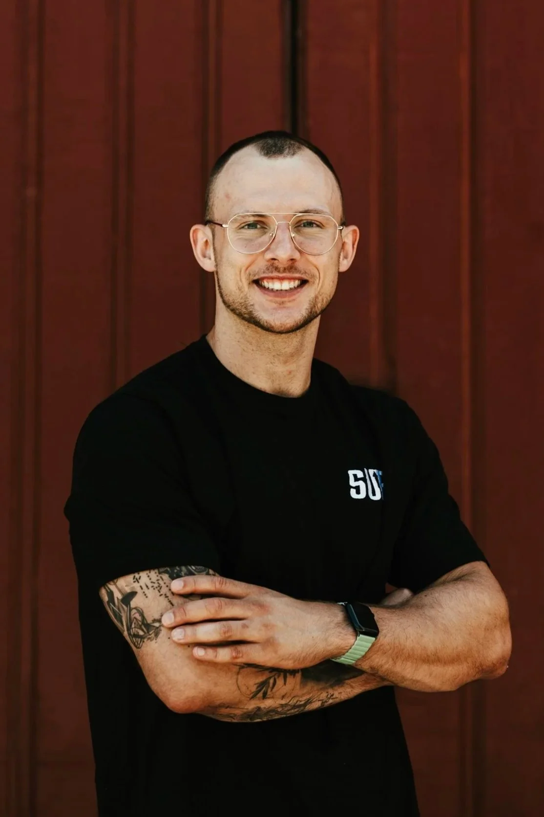 Coach Jonas von Straight UP steht in einem schwarzem T-Shirt vor einem roten Holzhintergrund steht, lächelnd mit verschränkten Armen.