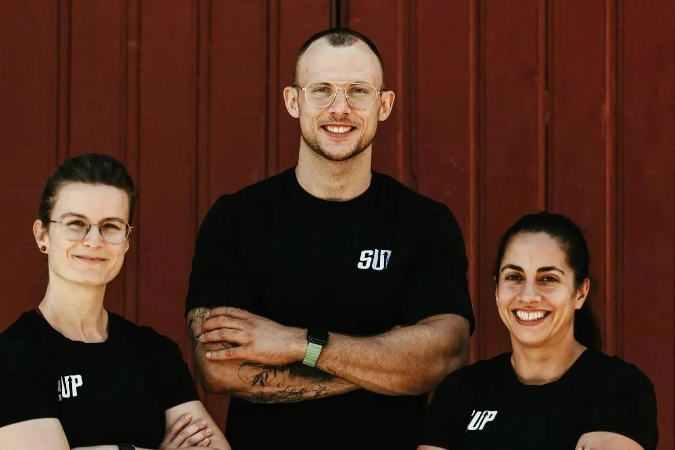 Drei smiling Menschen mit schwarzen T-Shirts, die vor einer roten Holzwand stehen, die den Hintergrund bildet.