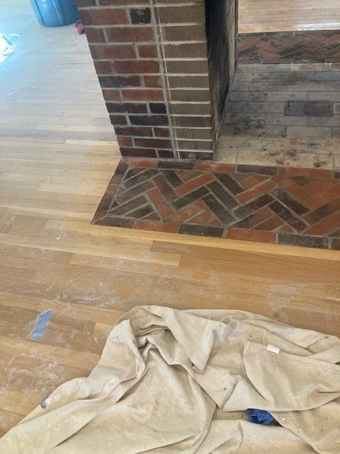 Close-up of a brick fireplace with a wooden floor and a section of patterned brickwork around the hearth, with a cloth on the floor.