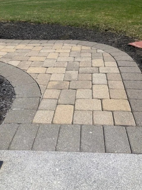 A circular pattern of gray paving stones surrounded by a curved border of light and dark gray bricks, with grass and dark soil at the edges.