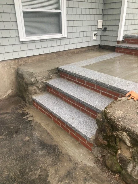 Newly constructed concrete stairs with brick trim leading up to a house porch, with a rock and dirt surrounding the stairs.