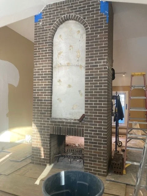 Brick fireplace structure with an arched opening and a firebox at the bottom, inside a room under construction.