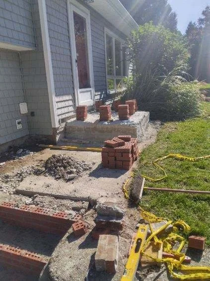 Construction site with bricks, a yellow framing square, and tools outside a house.