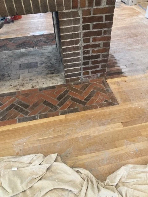 Interior scene showing a brick wall, a doorway with a brick and tile threshold, and a hardwood floor with a beige cloth in the foreground.
