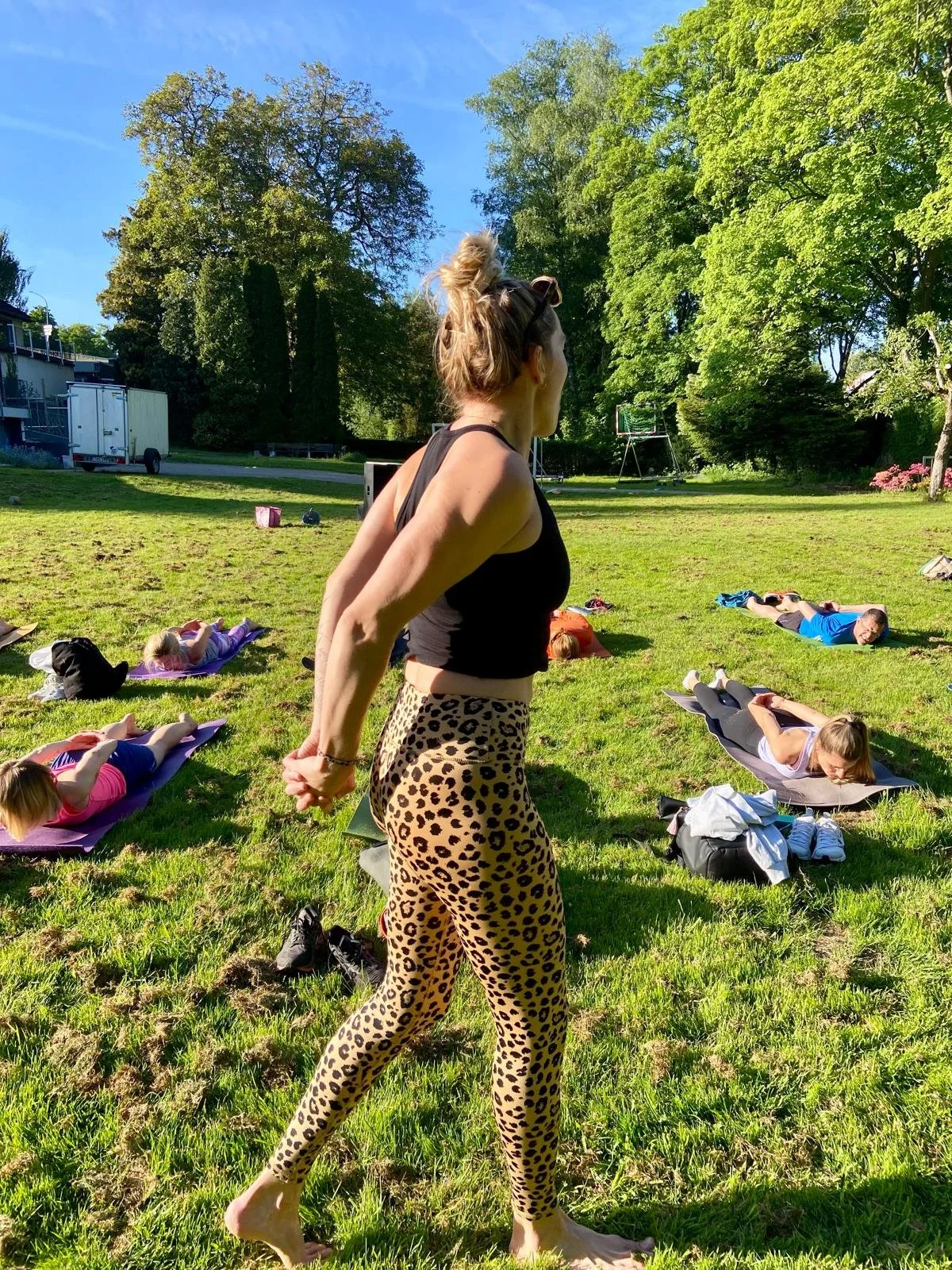 Eine Frau steht bei einem Yoga- oder Meditationskurs im Freien, während mehrere Personen auf Yogamatten liegen und strecken, umgeben von Bäumen und grünem Gras.