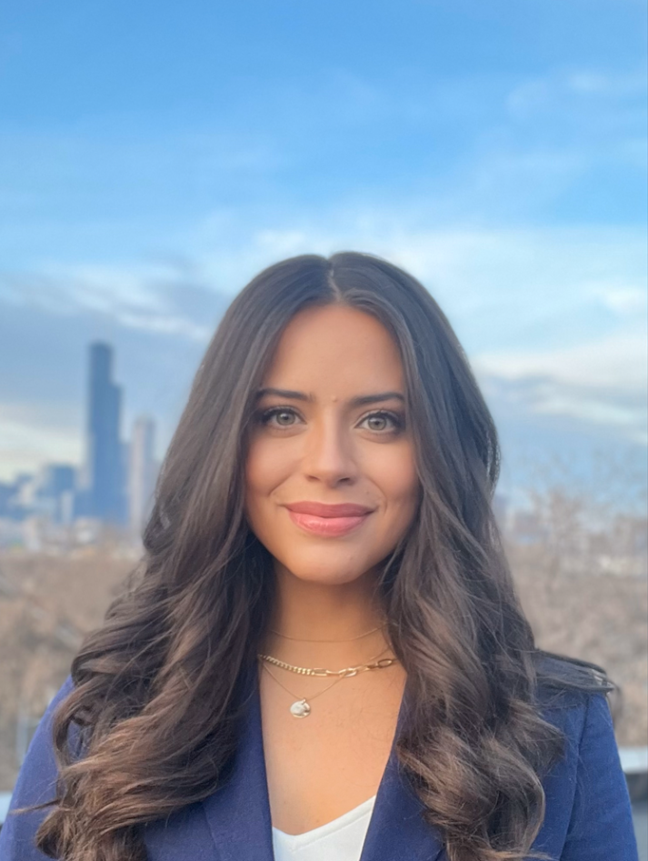 A woman with long wavy dark hair, wearing a navy blazer, white top, and gold necklaces, standing outdoors with a city skyline and blue sky in the background.