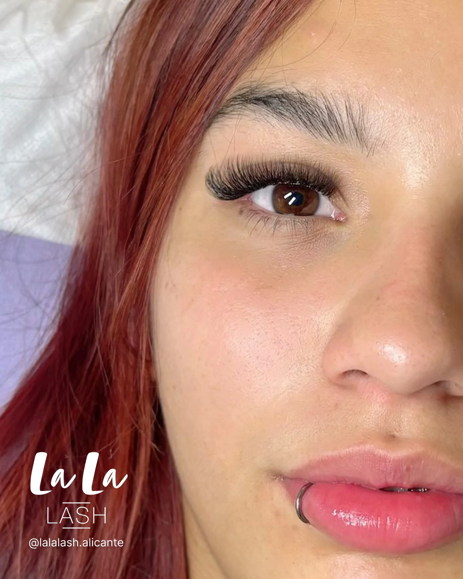Primer plano del ojo de una mujer con pestañas largas y rizadas, cabello de color rojo y parte de su mejilla visible, usando lápiz labial rosa y con un aro en la nariz.