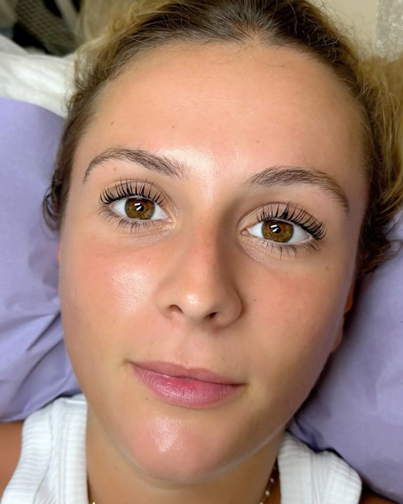 Primer plano del rostro de una mujer con ojos marrones y pestañas largas, con labios rosados y cejas bien definidas, usando una blusa blanca con rayas.