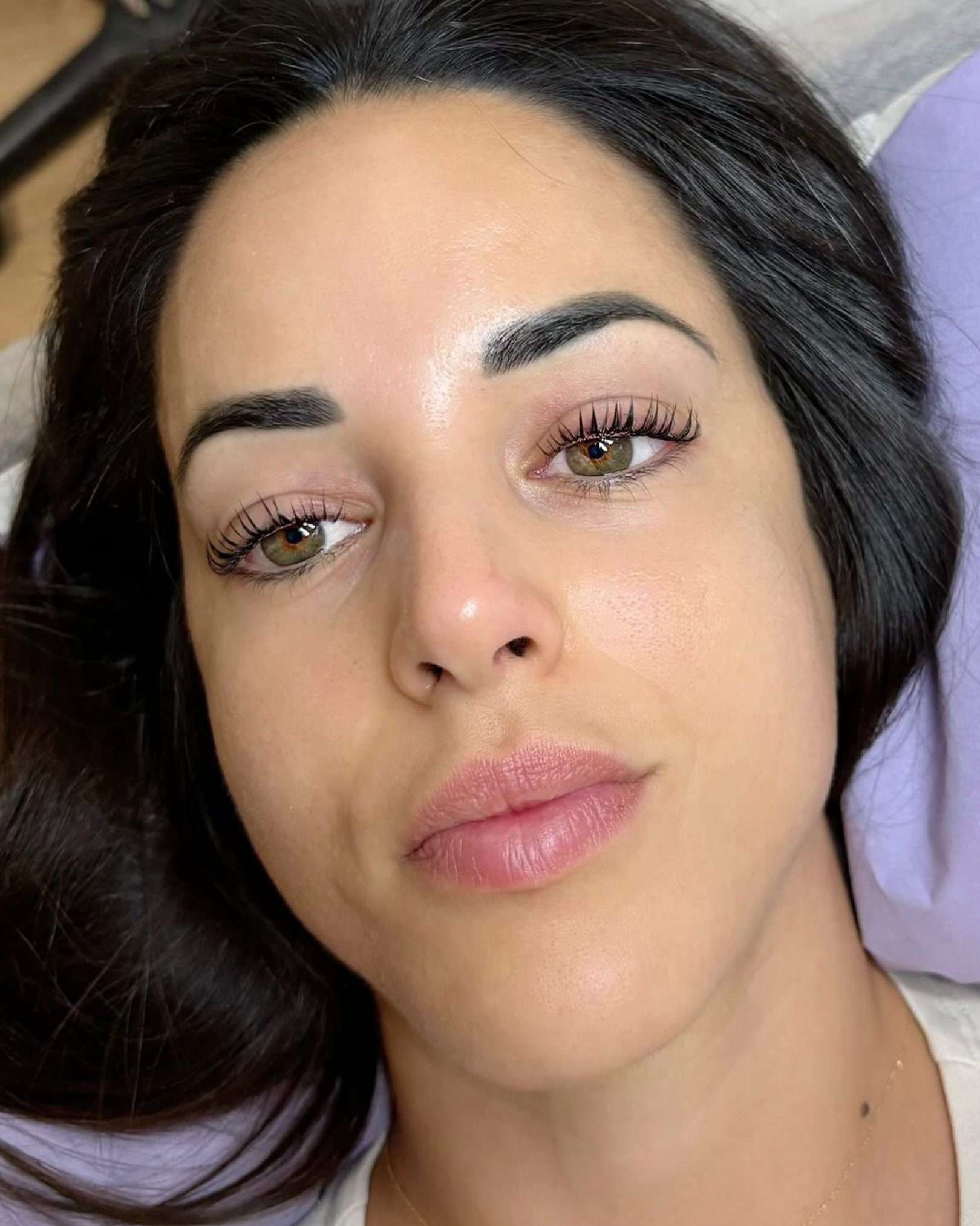 Primer plano de una mujer con cabello oscuro, cejas arregladas, ojos verdes con pestañas largas y maquillaje suave, con piel clara y labios naturales.