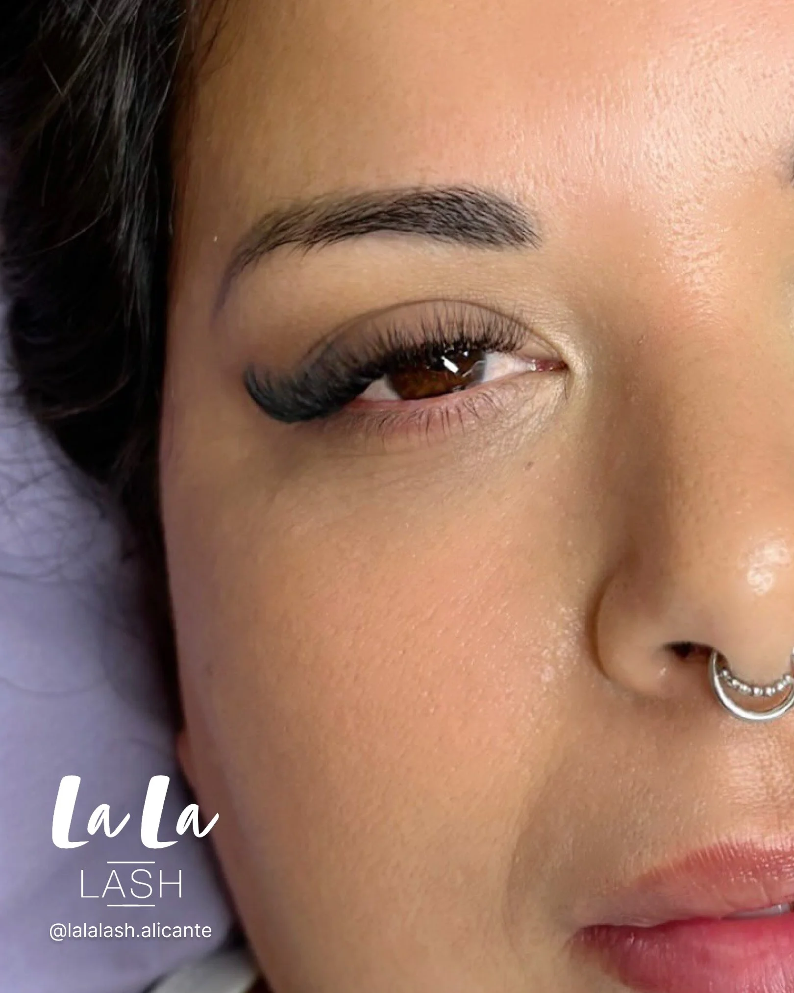 Primer plano de un rostro con cejas arregladas, maquillaje de ojos con pestañas largas y rizado, y labios suaves, con un piercing en la nariz. Texto en la imagen que dice 'La La' y 'Lash' con la cuenta de Instagram '@lalalash.alicante'.