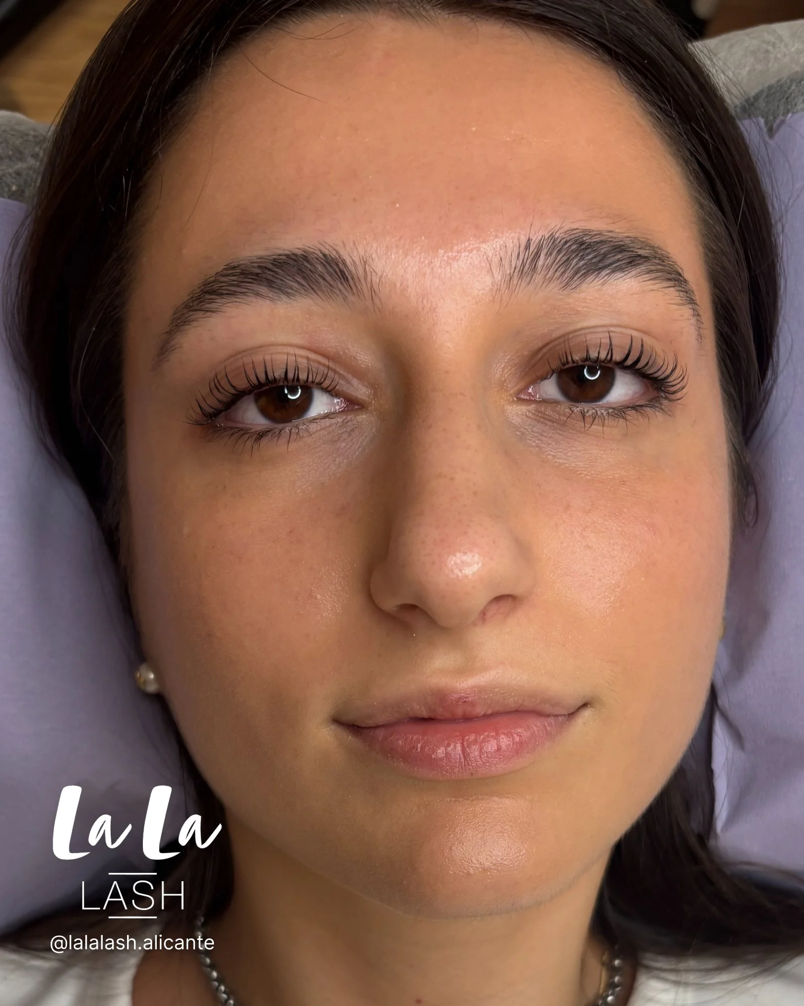 Primer plano del rostro de una mujer con cejas arregladas y pestañas largas, ojos marrones y labios rosados. Tiene piel clara y cabello oscuro. Se aprecia una parte del cuello con collar de perlas y una blusa blanca.