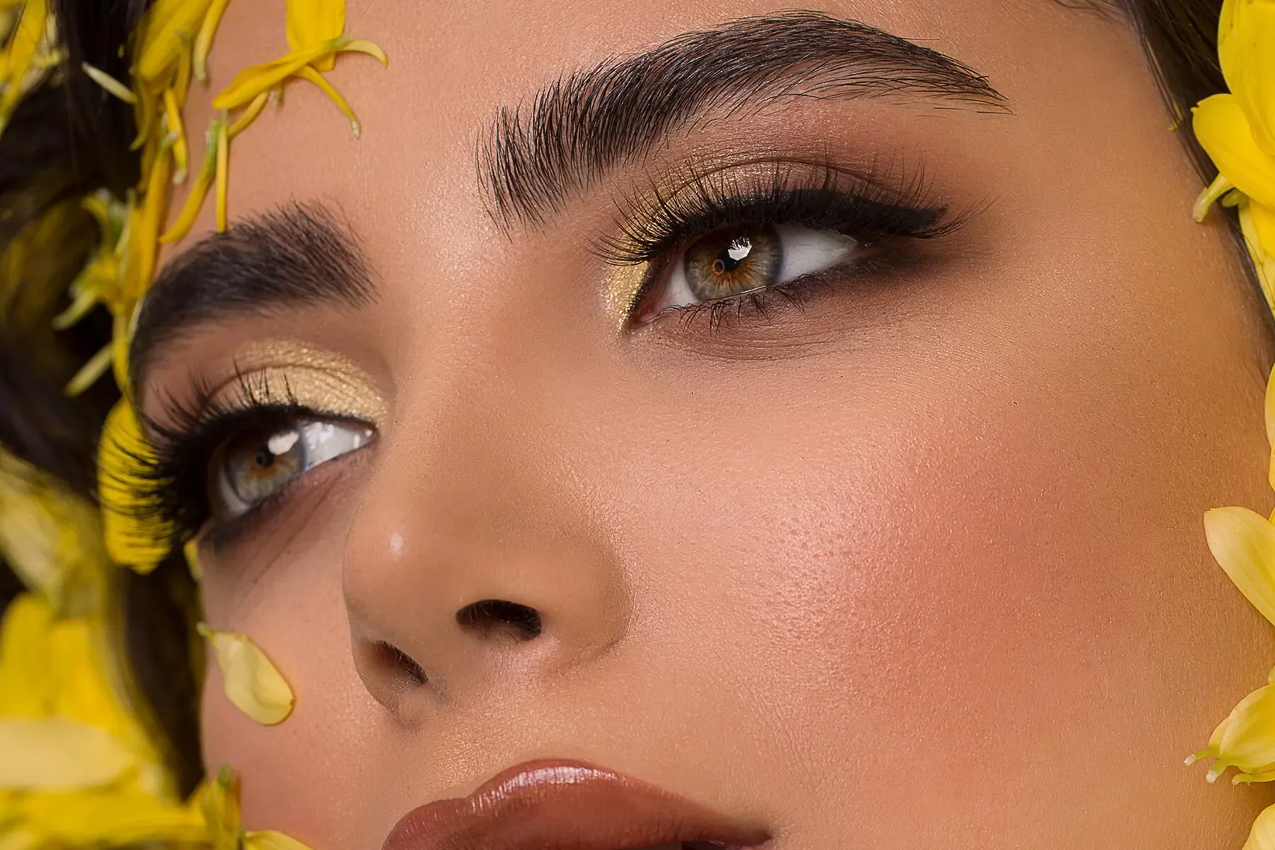 Primer plano del rostro de una mujer con maquillaje y cejas arregladas, rodeada de flores amarillas.