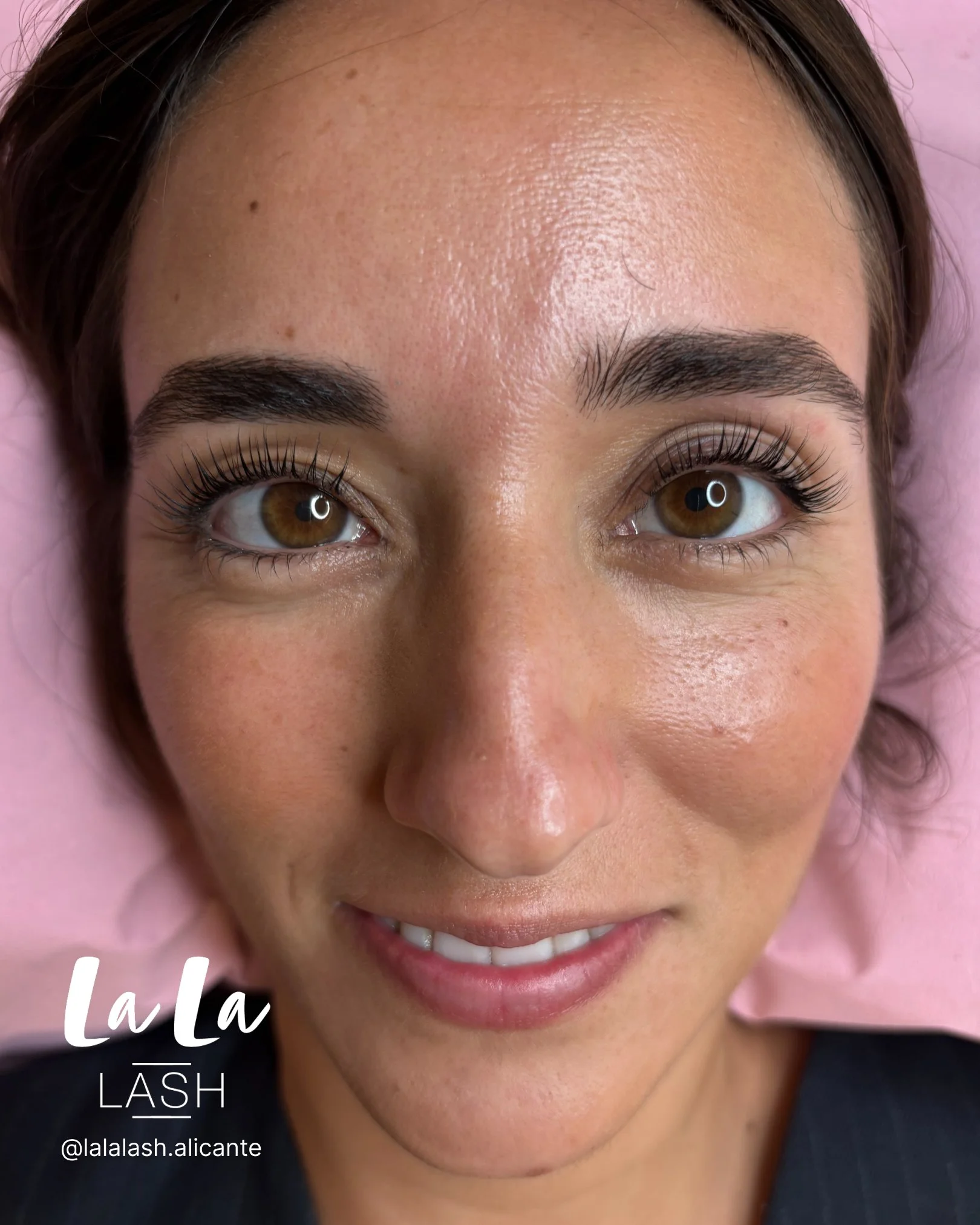 Retrato de rostro de mujer con cejas arregladas y pestañas largas, sonriendo ligeramente, con piel brillante y ojos marrones, en un entorno de salón de belleza.