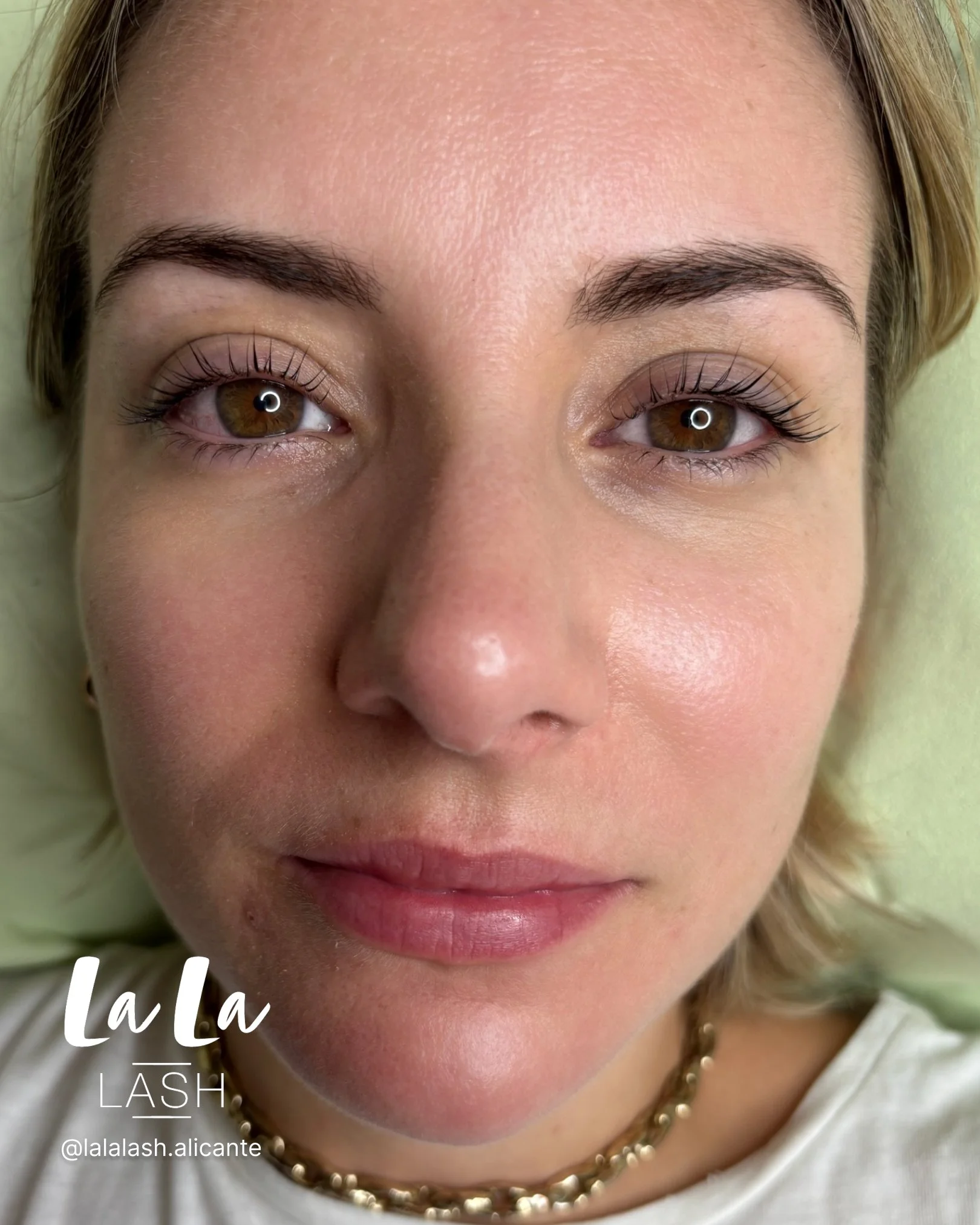 Primer plano de rostro femenino con pestañas postizas, cejas definidas y labios pintados de rosa, usando una cadena de perlas y ropa blanca, con fondo borroso en tonos verdes.