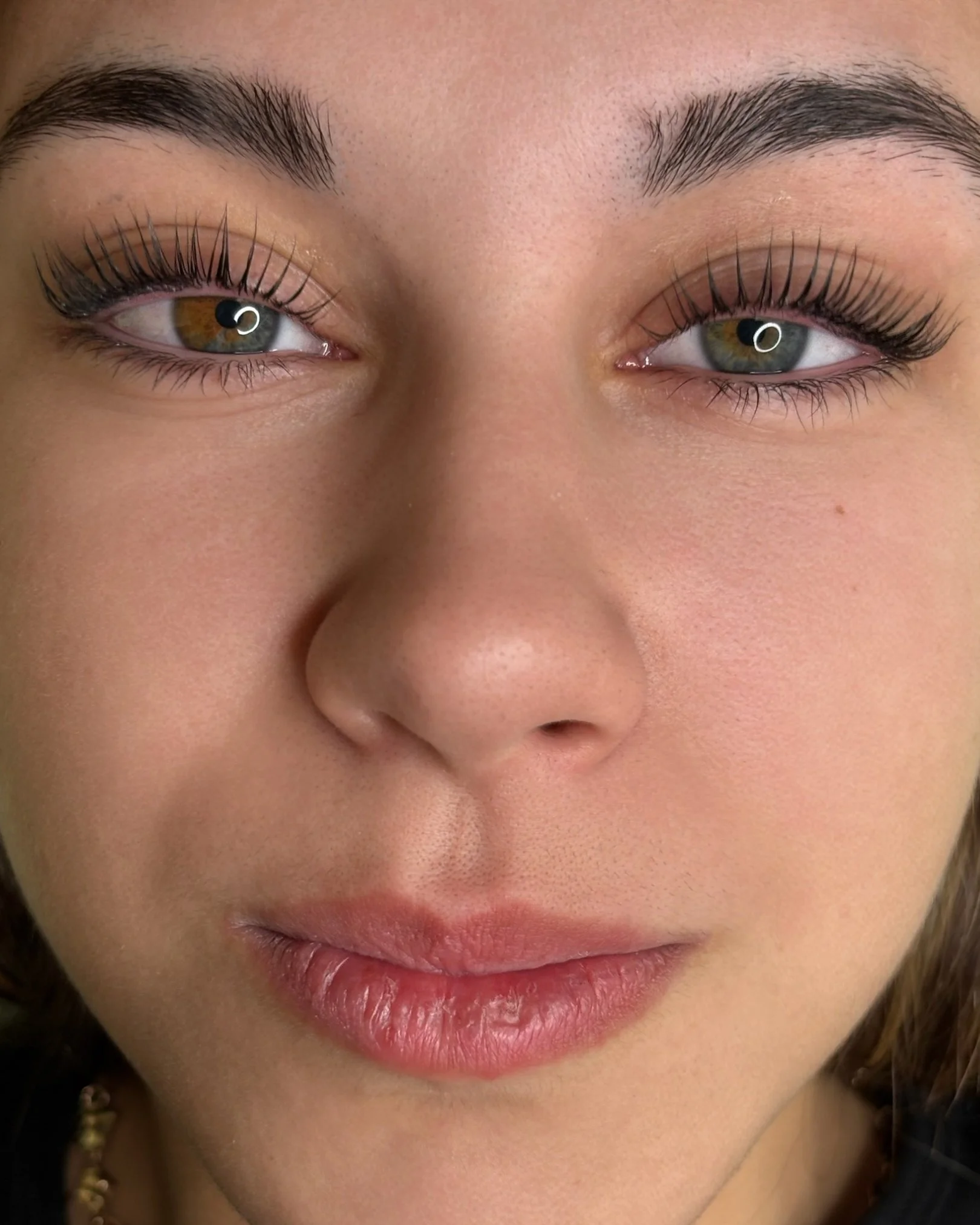 Primer plano del rostro de una mujer con cejas arregladas, pestañas largas y delineadas, piel clara, labios sonrojados y ojos de color gris con lentes de contacto de color ambar.