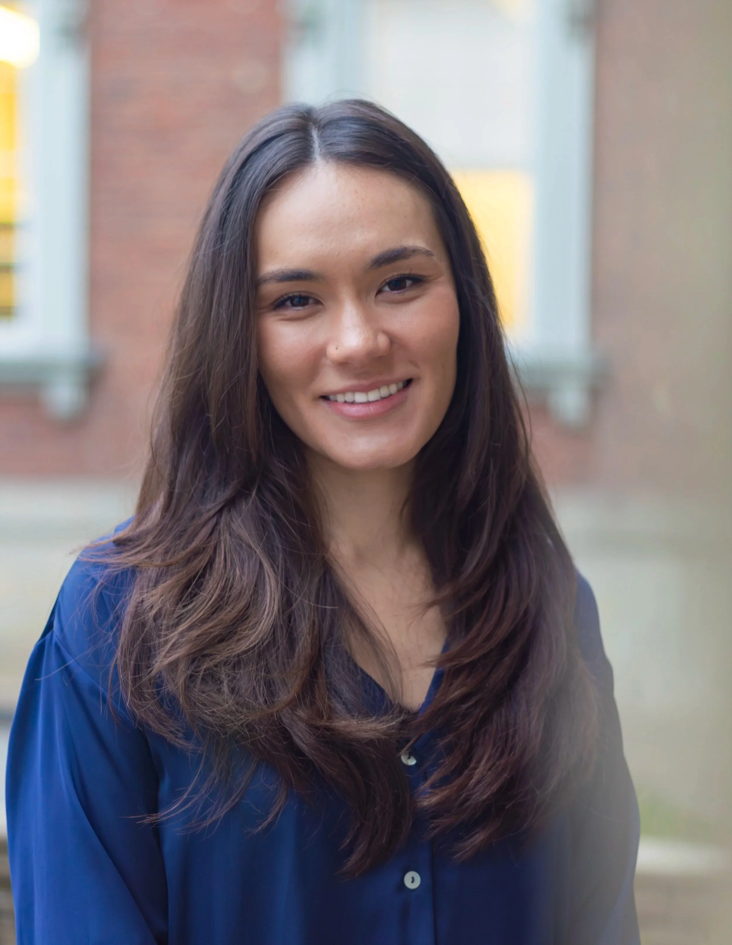 Close-up of a young woman with long brown hair wearing a blue shirt, smiling outdoors with blurred background.