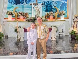 A young boy and man standing together in front of an altar decorated with flowers, statues, and a painting of the Virgin Mary, inside a church.