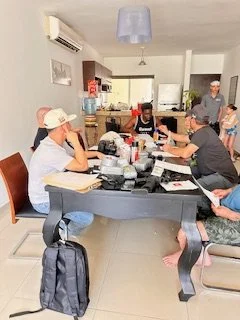 People sitting at a table in a kitchen, engaging in conversation or work, with various items on the table.