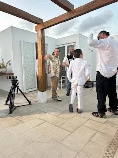 Group of five people standing on a terrace with white and glass walls, some dressed in formal attire, and a camera on a tripod. Overcast sky visible.