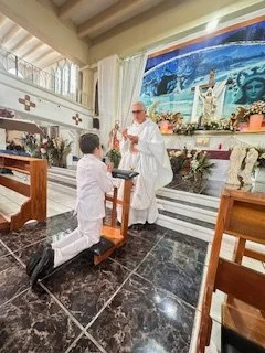 A clergy member performing a religious ceremony in a church, with a kneeling boy in white robes and another boy standing with hands clasped in prayer.