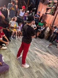 A young boy in black shirt and red pants dancing in front of a group of children and adults in an indoor space with wooden walls.