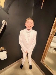A young boy in a white tuxedo and bow tie standing indoors, smiling at the camera.