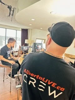A man in a black t-shirt and hat, with 'AlbertaLIVE.ca CREW' written on the back, is in a studio setting with multiple monitors and another person sitting at a desk, possibly filming or recording a video.