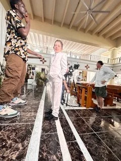 A woman in a white outfit standing in a church or chapel with wooden pews, two young boys nearby, and a man in white behind them. The woman is smiling and facing the camera.